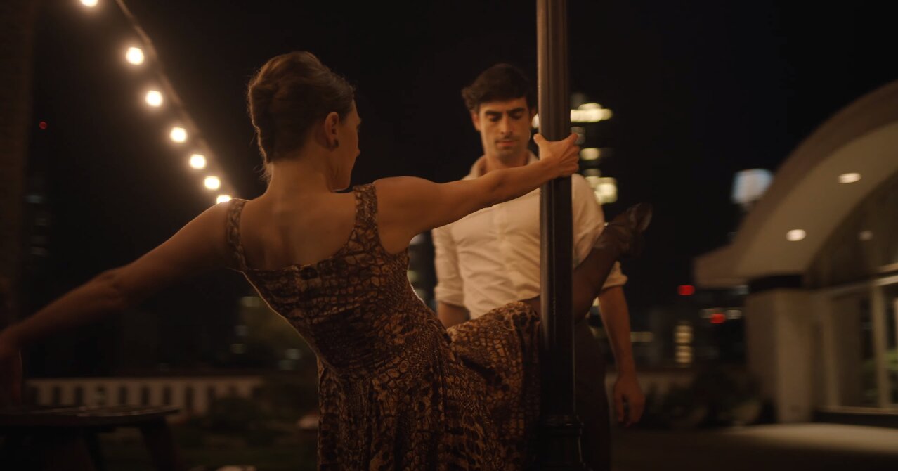 Two dancers in a dramatic pose around a metal pole on a night terrace with string lights, from the Mrs Robinson short film.